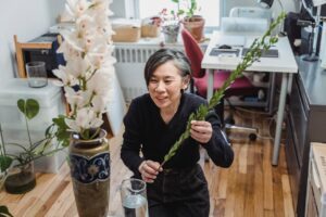An Asian woman creating a flower arrangement in a cozy, indoor home office setting, surrounded by plants.